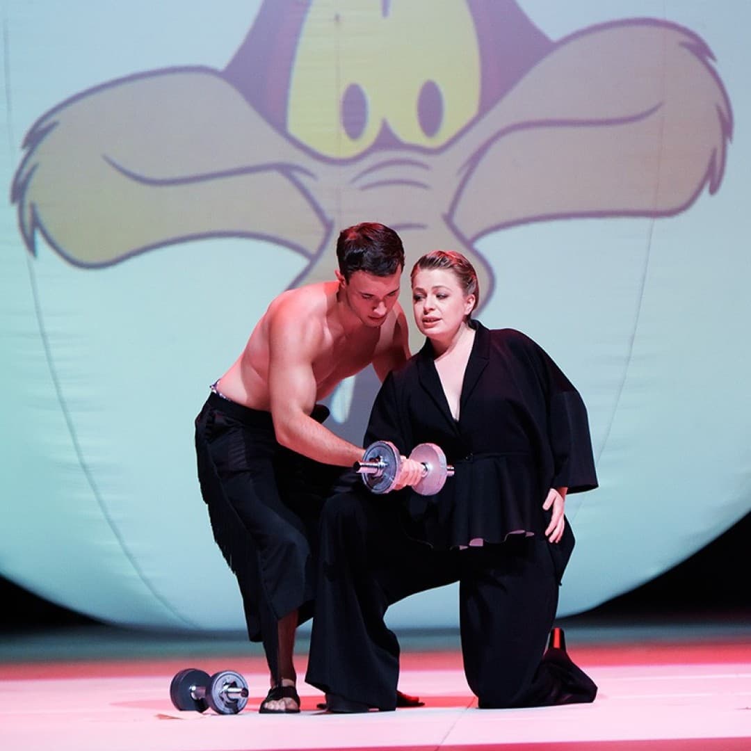 Two people with a dumbbell; one helps the other lift. Background: large projection of Wile E. Coyote's face.