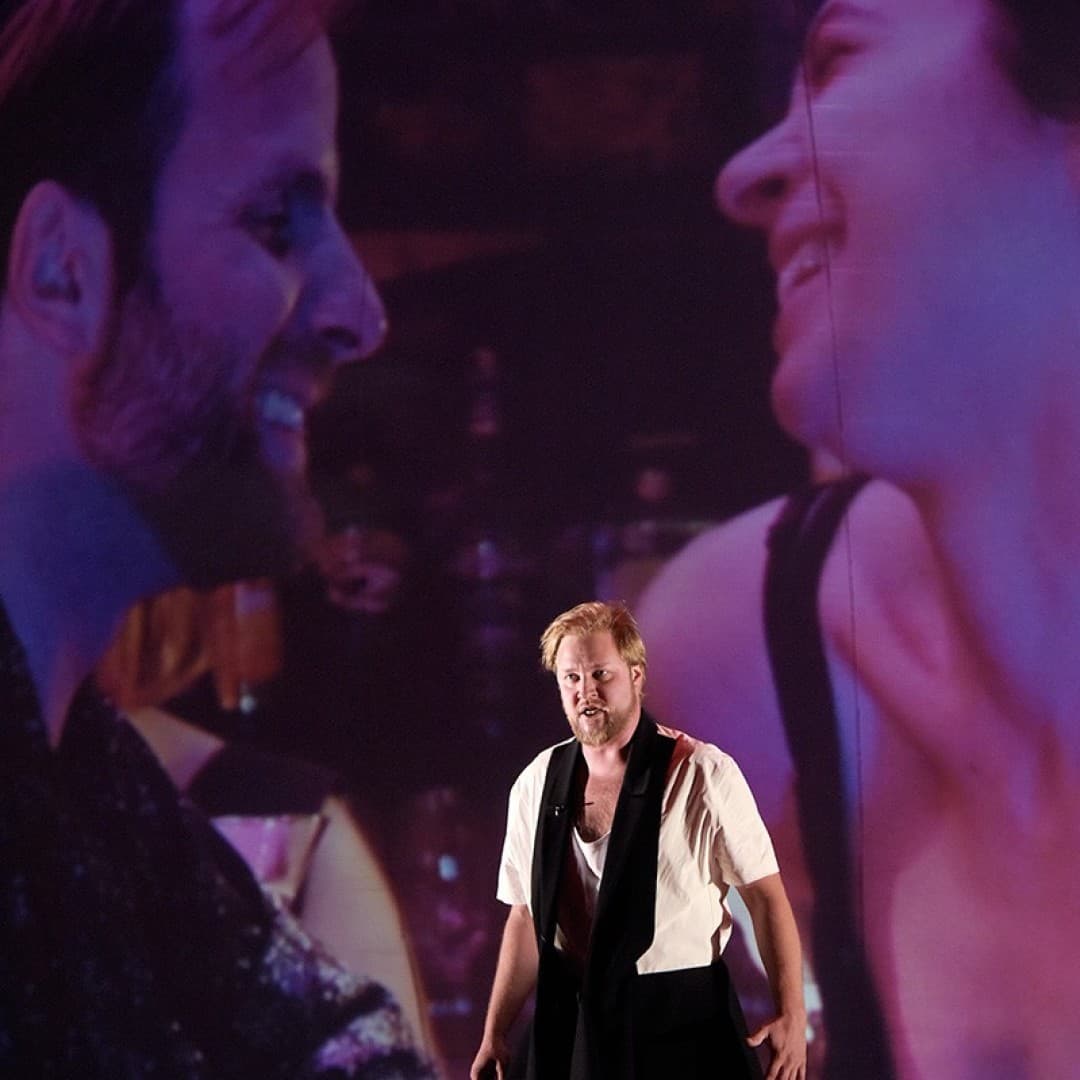 A person stands in front of a large projected couple. The person wears a white shirt, dark pants, and scarf. The couple is smiling at each other.