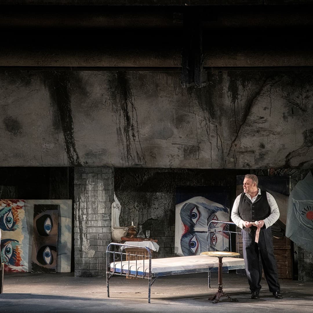 Two people are in a dimly lit room with large eye paintings on walls, a metal bed, and a small table. The person on the left is seated, the right is standing.
