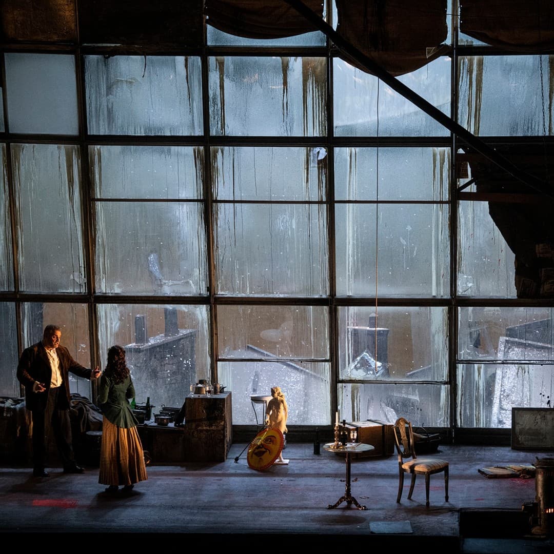 Two people facing each other on a stage with a large industrial window and minimal furniture, including chairs and a table, in the background.