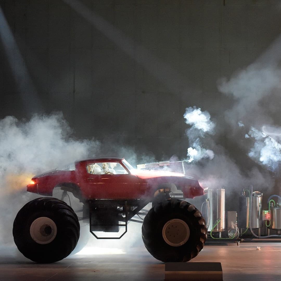 A red monster truck on stage emits smoke. A person on the left takes a photo. Metal containers with hoses and lights are in the background.
