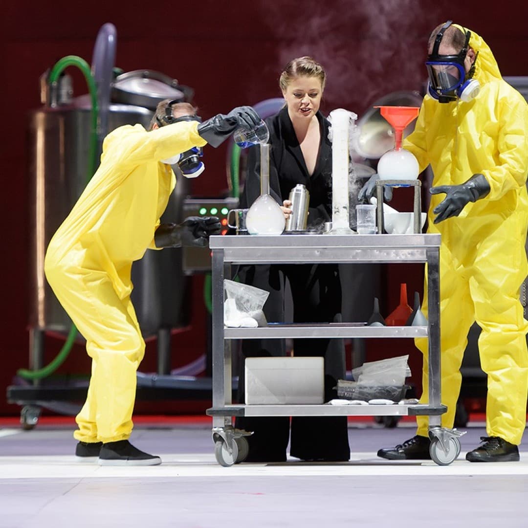 Three people in protective suits and a black outfit stand around a table with steaming containers in a laboratory setting.