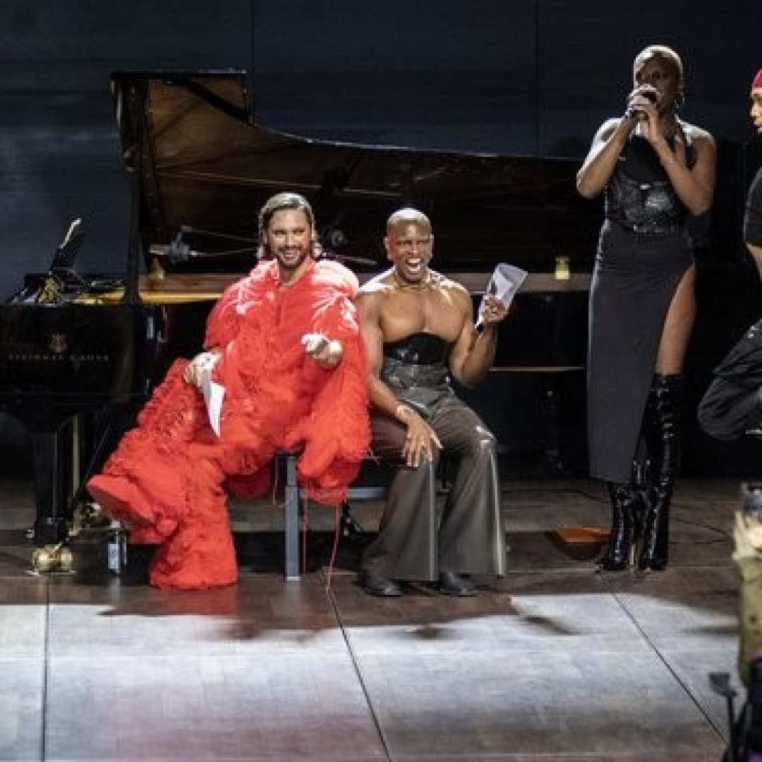Three seated performers with vibrant outfits on stage, two others dance energetically; grand piano in the background, audience visible in foreground.