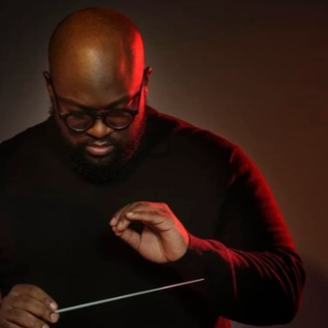 Person in dark clothing holding a conductor's baton, looking downward, with a dark background and red lighting on one side.
