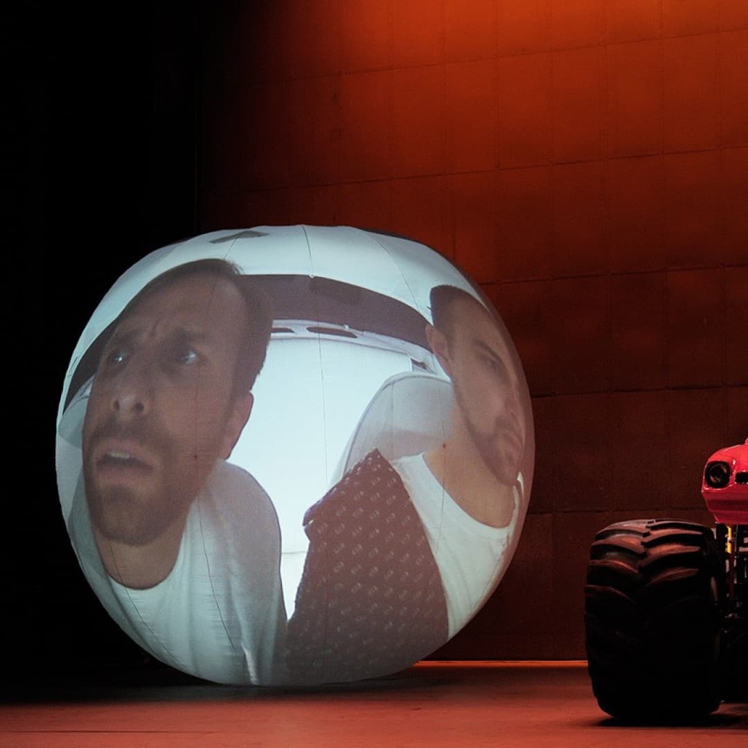 Two large faces projected on a spherical screen beside a red monster truck with two people inside, against a dimly lit orange background.