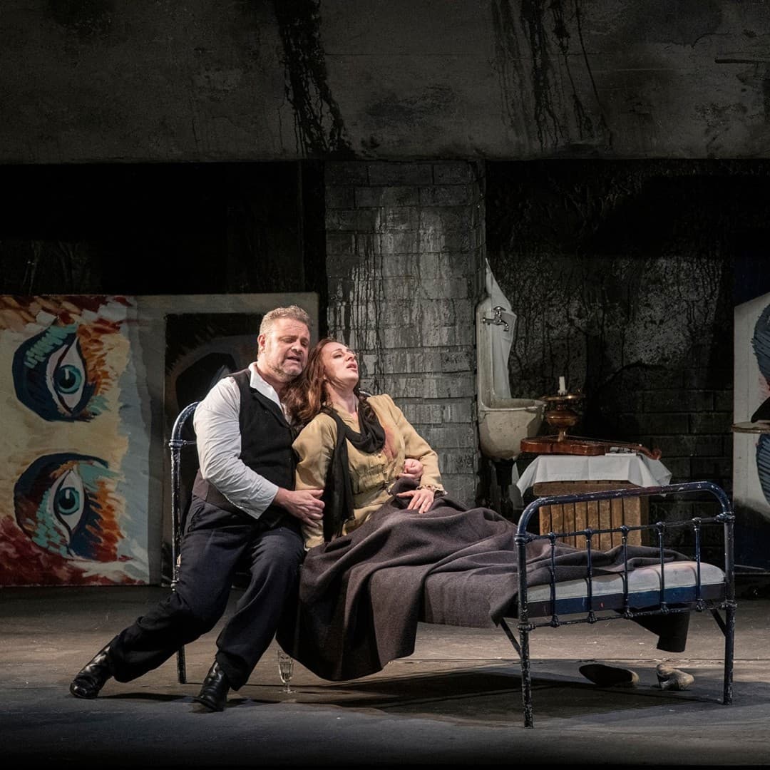 Two people lean on a metal bed, one holding the other. Another person in a hat and long dress stands nearby. Large eye paintings decorate the walls.