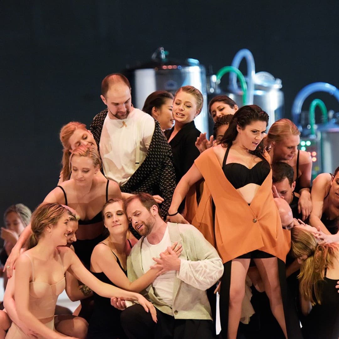 Group of performers in a dance formation, several embracing. Some wear black or flesh-toned outfits. Industrial equipment visible in the background.