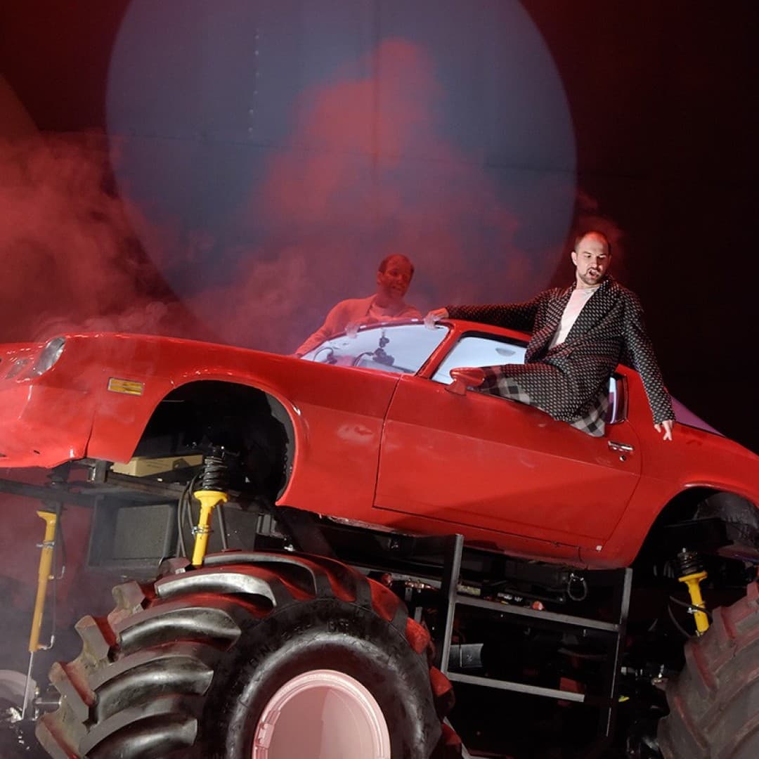 Two people in red car raised on large tires, one standing through sunroof, spotlight and smoke around, person in checkered suit sitting on car window ledge.
