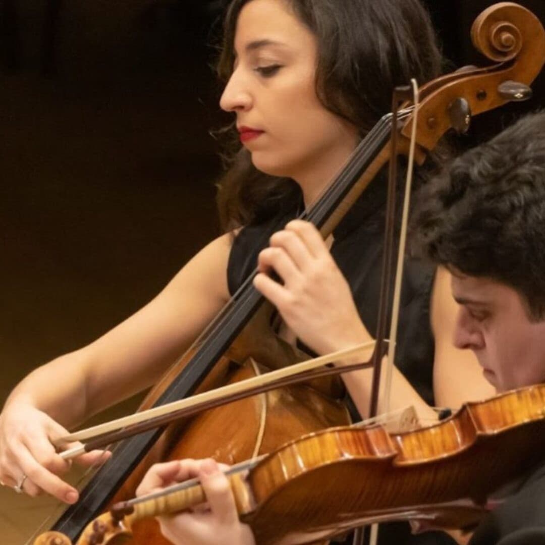 Person playing cello with focus, another person playing viola beside, both in formal wear, on a dimly lit stage.