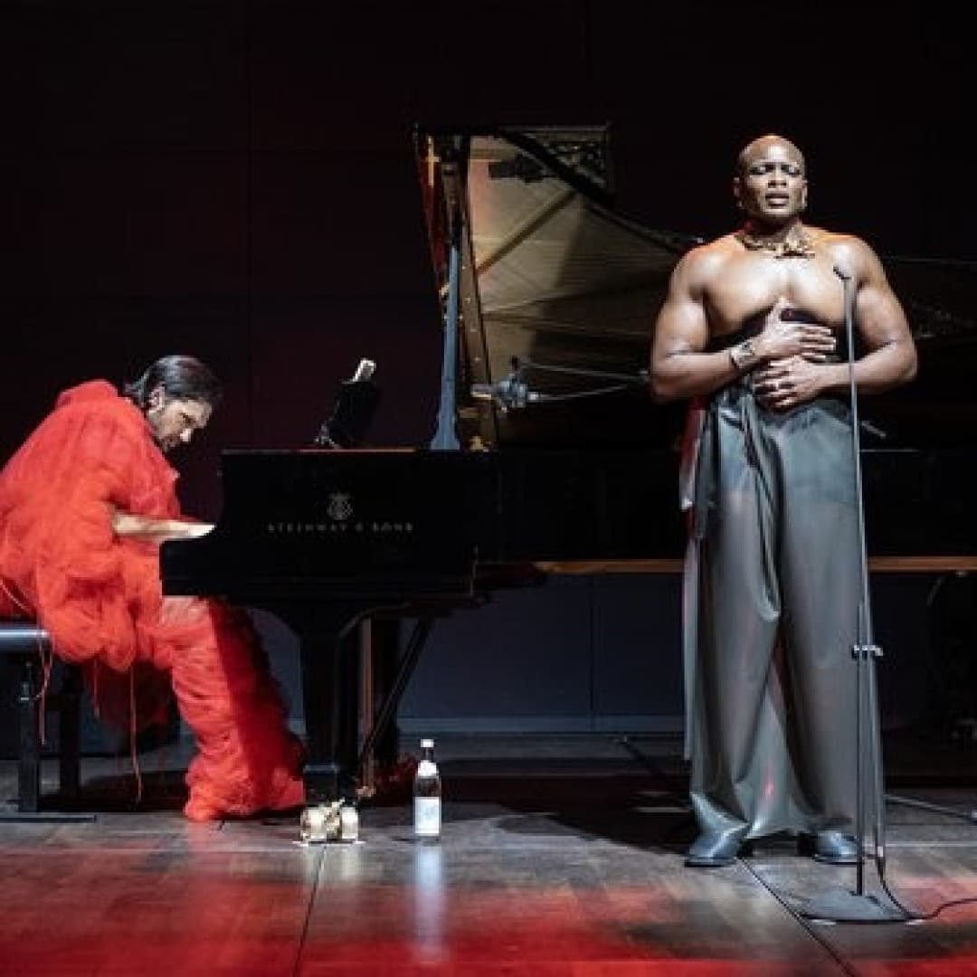 Person in red playing the piano. Another person sings into a microphone, wearing wide gray pants. A bottle is on the floor. Stage is dimly lit.