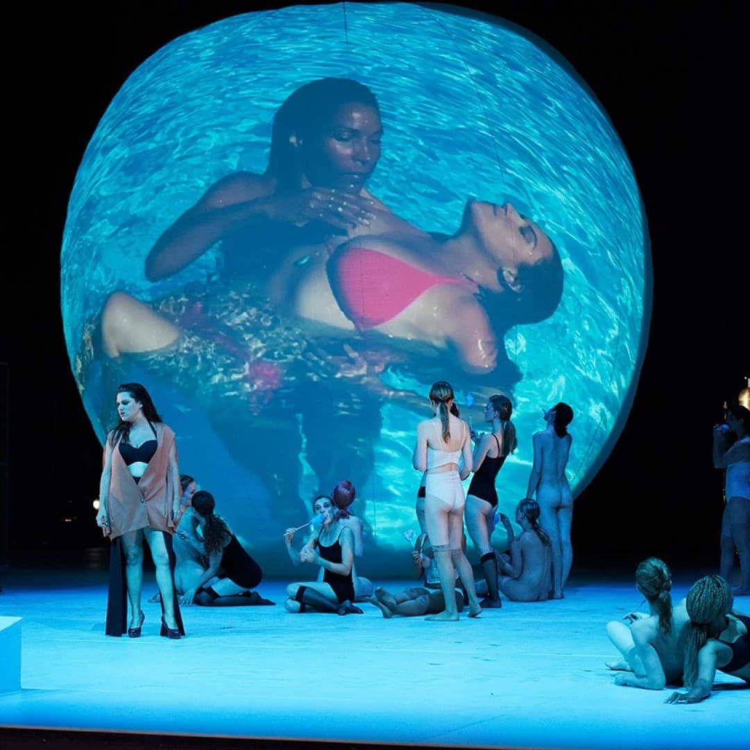 Large projected image of two people underwater in a pool. Several people in swimwear on stage, one holding blue fabric with "DREAM" written on it.
