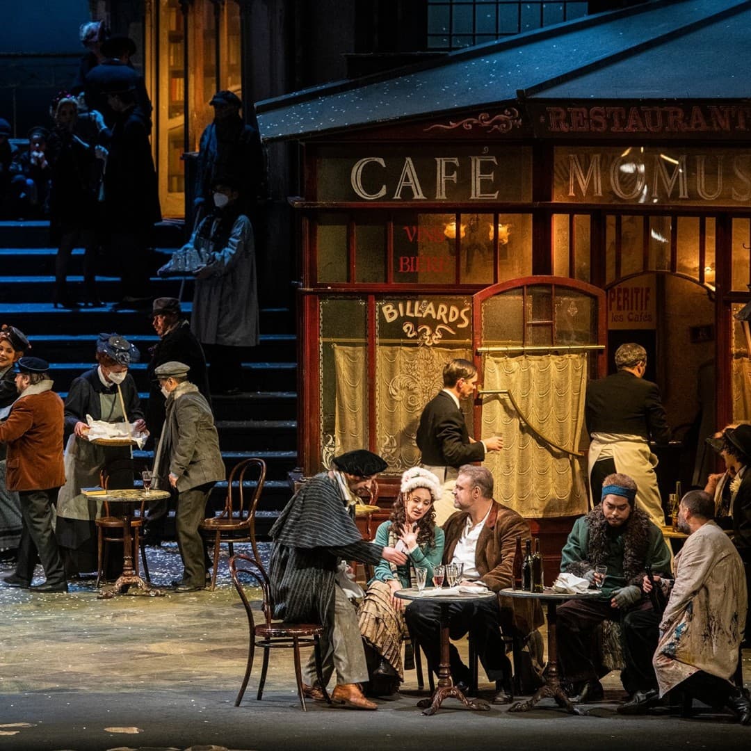 Multiple people gather in front of Cafe Momus, with tables, a person serving, and a red-dressed person sitting. Steps in the background with seated figures.