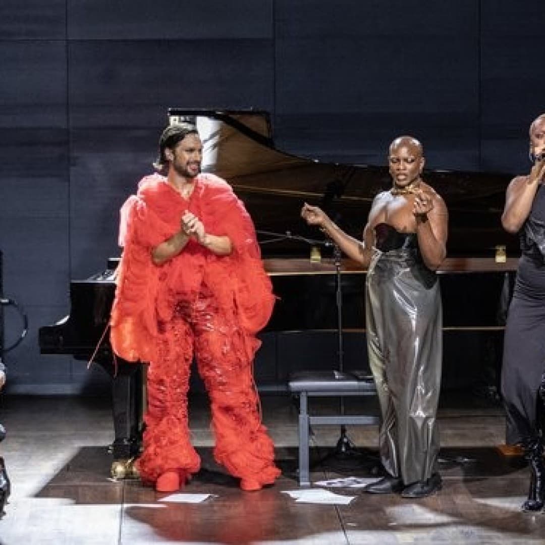 Four performers stand before a piano on stage. One wears a bright red outfit, while three wear shiny black attire. Audience members are seated nearby.