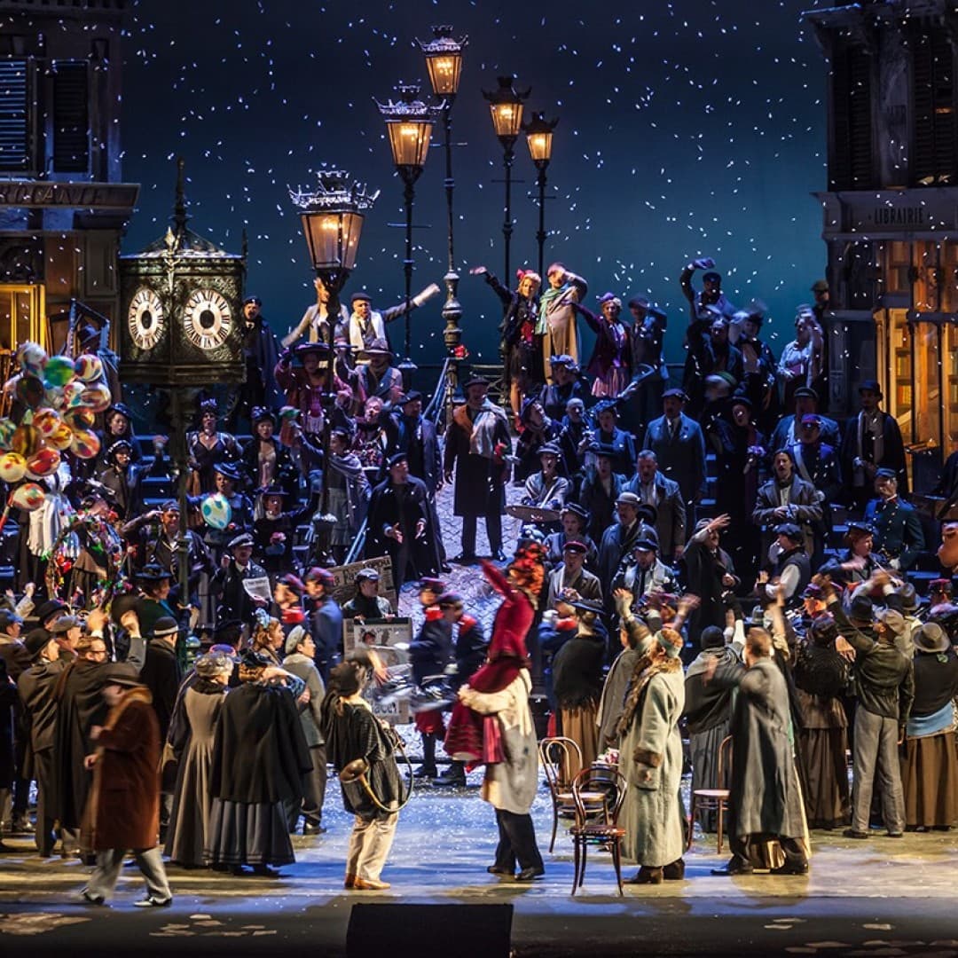 Crowded stage with people in winter clothes, colorful balloons, snow falling, streetlamps, and storefronts marked as Brigganti and Librairie.