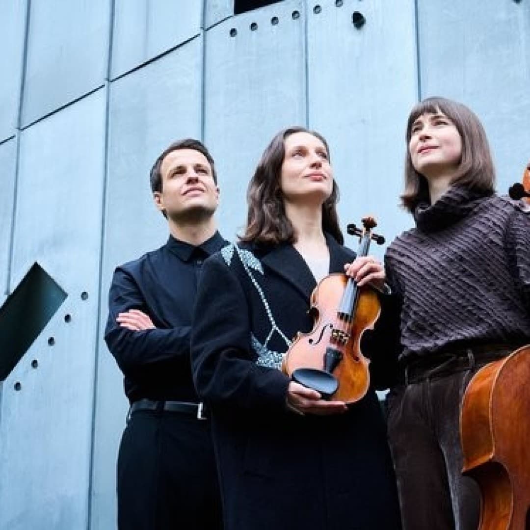Three people with string instruments stand in front of a large metal wall. Two hold violins, one holds a cello. They look upward.