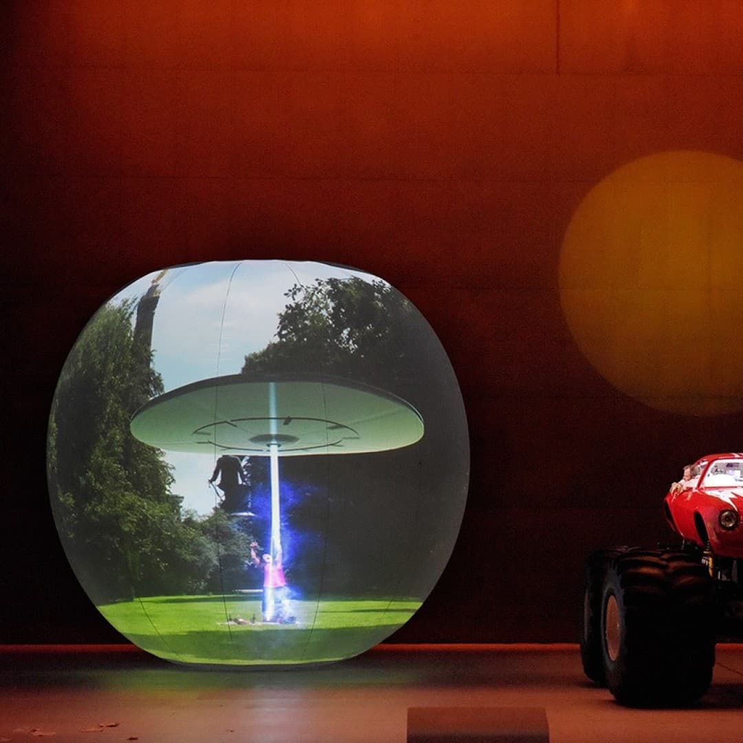 Large sphere with a UFO projection on left, red monster truck with a person standing beside it on right, under an orange-lit background.