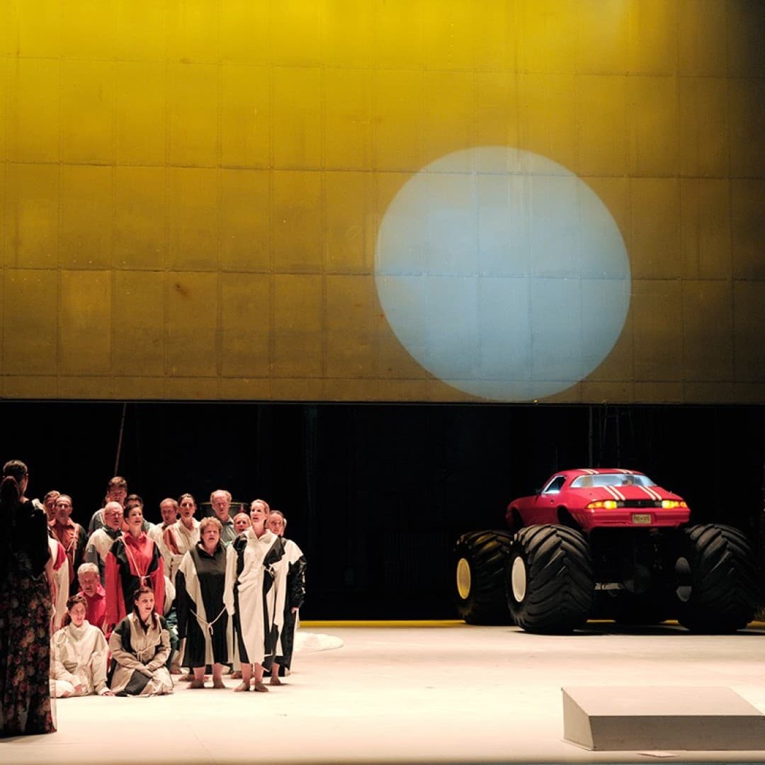 Group of people in robes stands on stage under yellow lighting; to the right, a large red monster truck with white stripes.