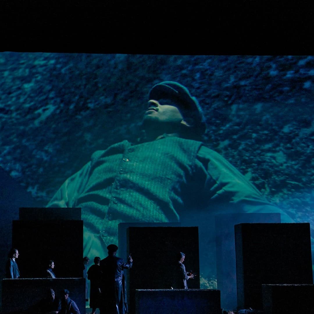 Large projection of person in cap and vest on stage backdrop, multiple silhouettes visible in front of the screen, seated person on the left.