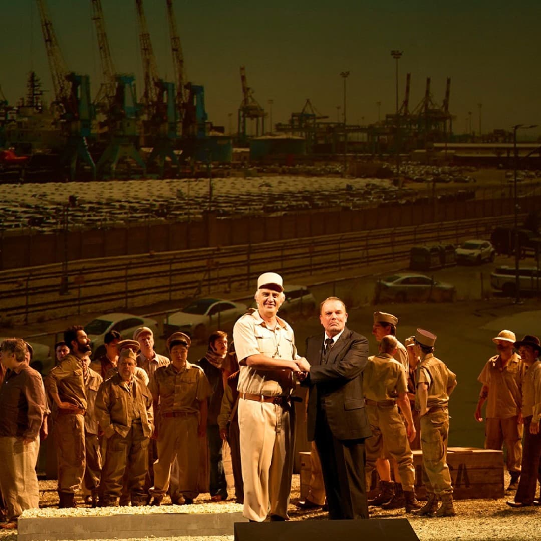 Two people shake hands center stage; one in military uniform, one in a suit. Group in beige attire surrounds them. Industrial port background with cranes.
