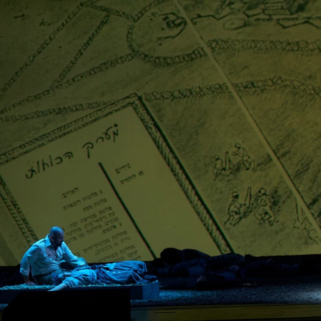 A person in a suit stands on stage; another person in white sits on the floor. A large background with Hebrew text and illustrations is visible.
