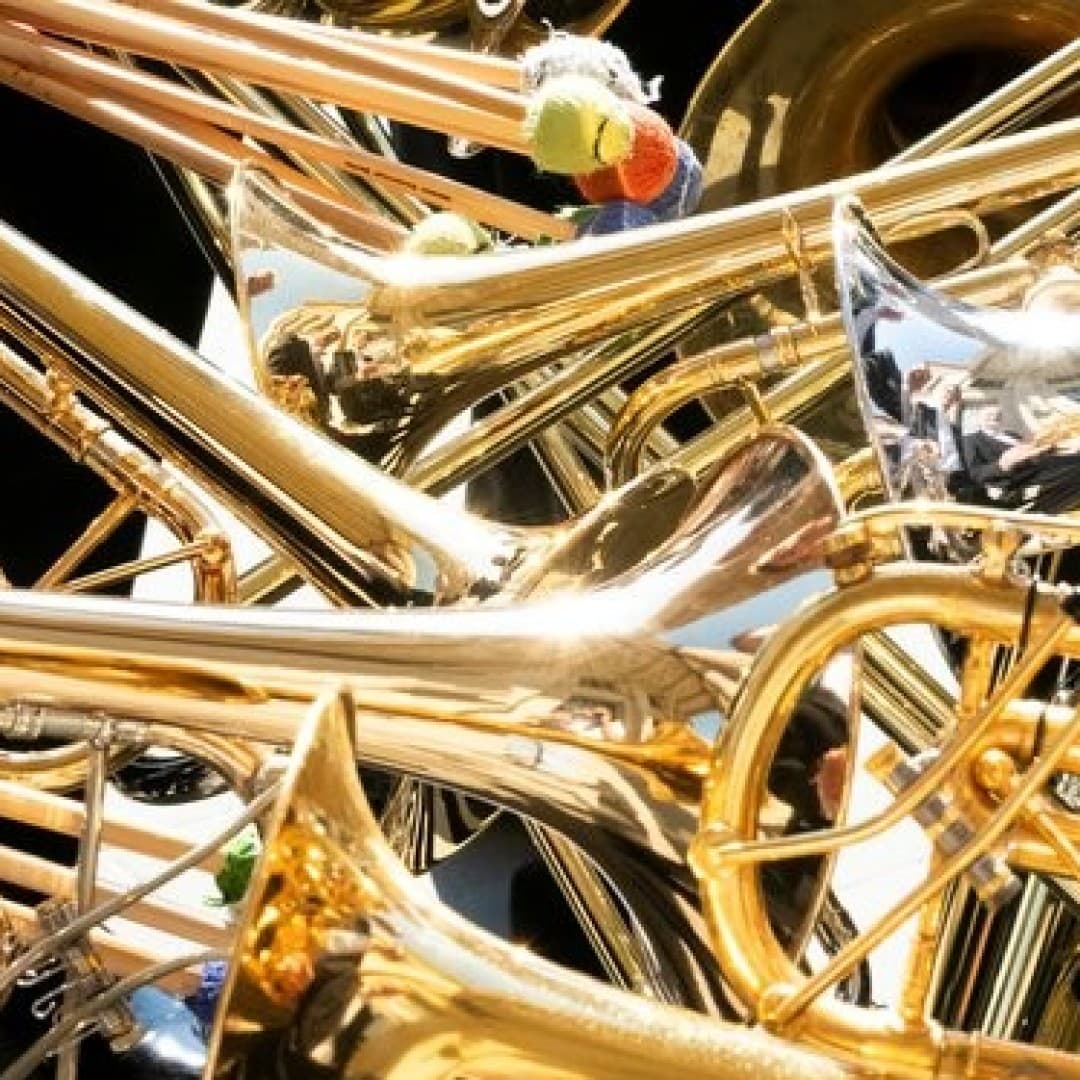 A jumble of overlapping brass and silver trumpets and trombones with drumsticks visible in the top left corner.