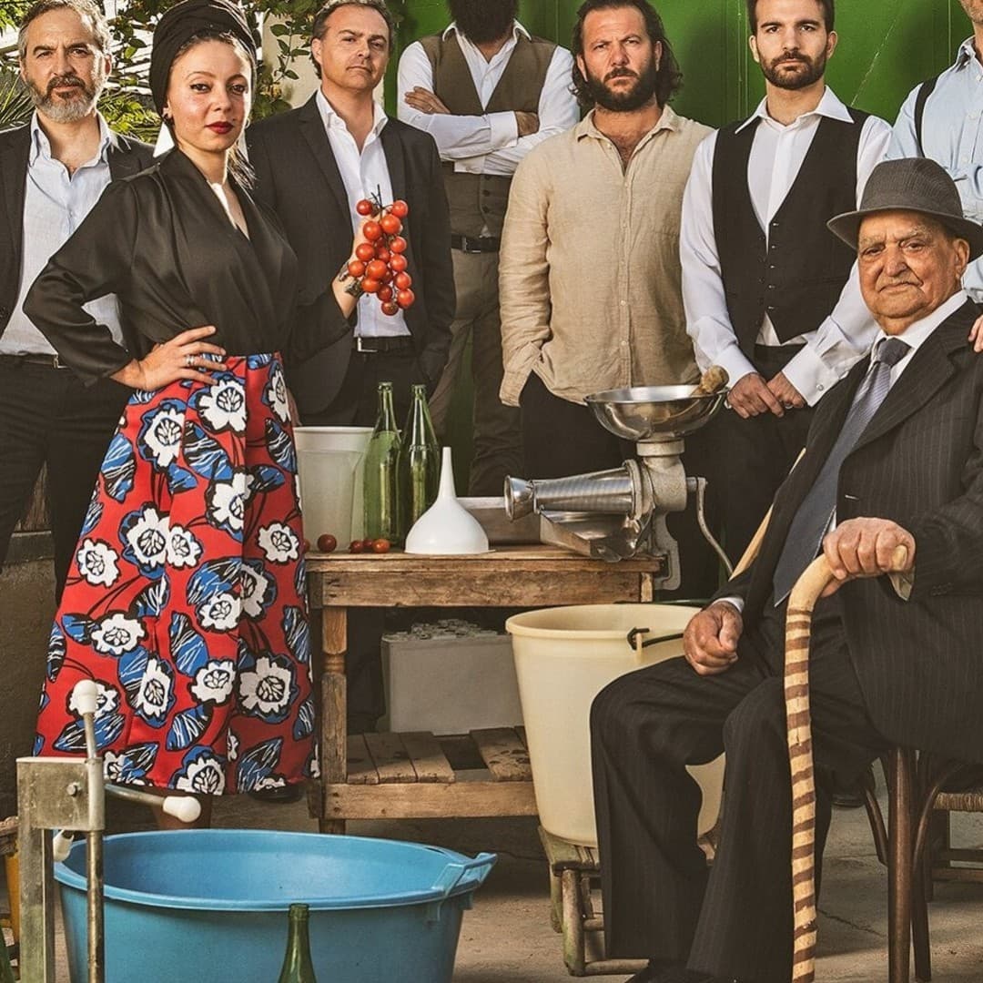 Group of people posing outdoors. One seated with a cane, a person holding tomatoes, and a pregnant person in a long dress. Bottles and crates nearby.