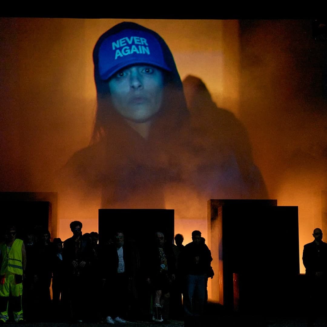 Large projected face wearing cap with text "NEVER AGAIN" above people standing in front of illuminated smoky background.