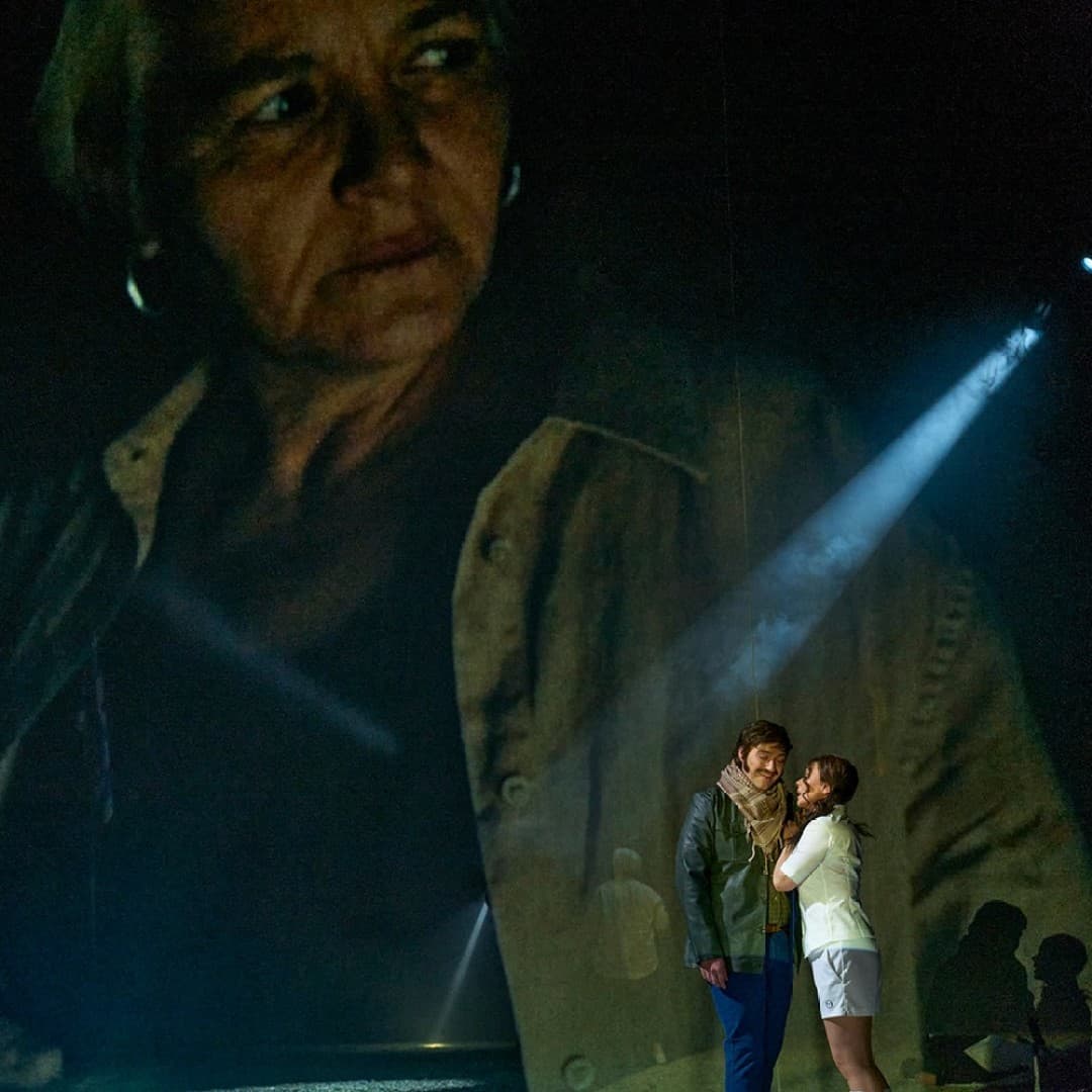 Large projection of a person with hoop earrings. In front, two people stand under a spotlight, one wearing a scarf, the other in white hugging.