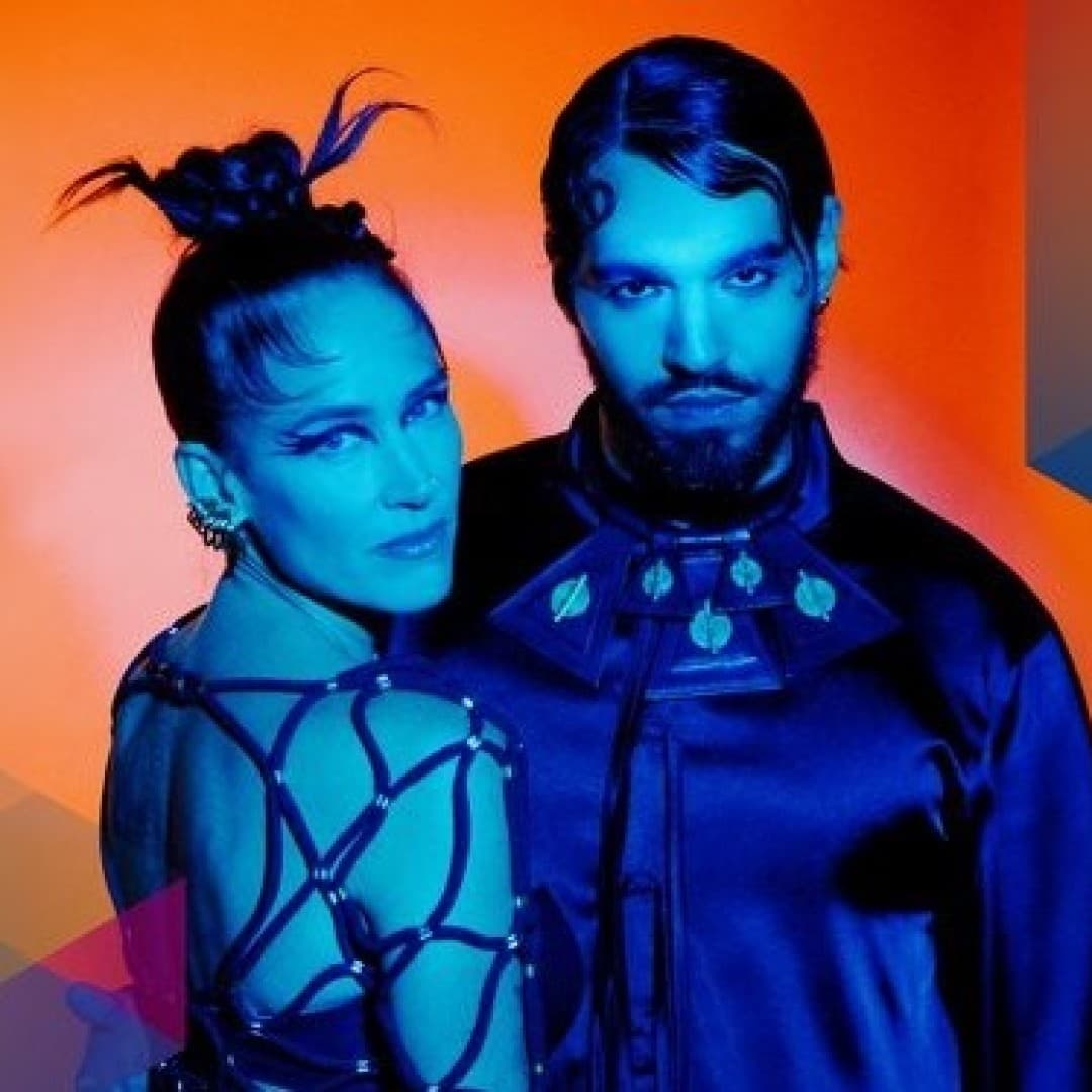 Two people standing closely; female-read person with a bun and harness top, male-read person with long hair and dark outfit; orange and blue geometric