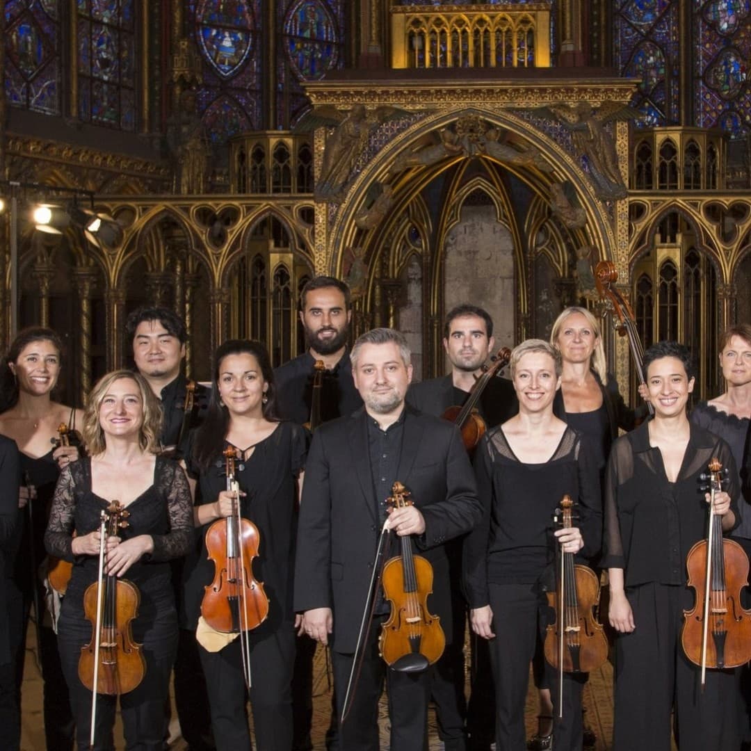 Thirteen people holding string instruments in a gothic church interior, all wearing black clothing, standing in front of ornate golden arches.