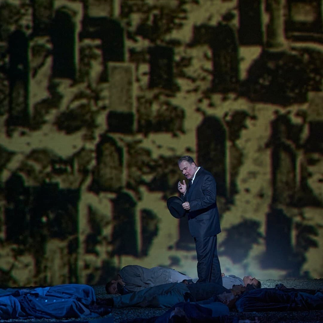 A person in a suit holds a hat, standing among lying figures. In the background, a projection of a cemetery with gravestones.