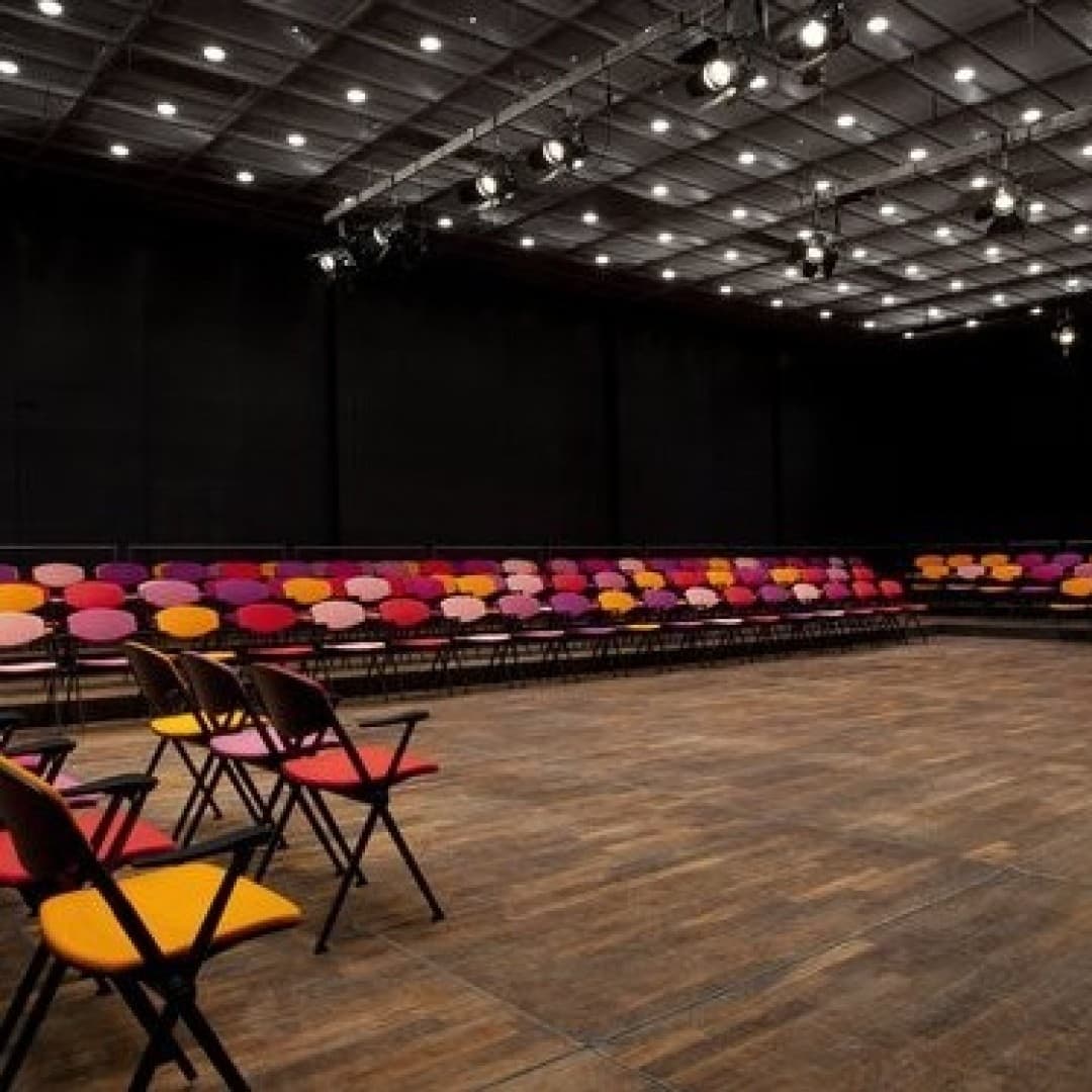 Spacious room with wooden floor, filled with rows of colorful folding chairs in pink, orange, and purple. Ceiling has multiple spotlights.