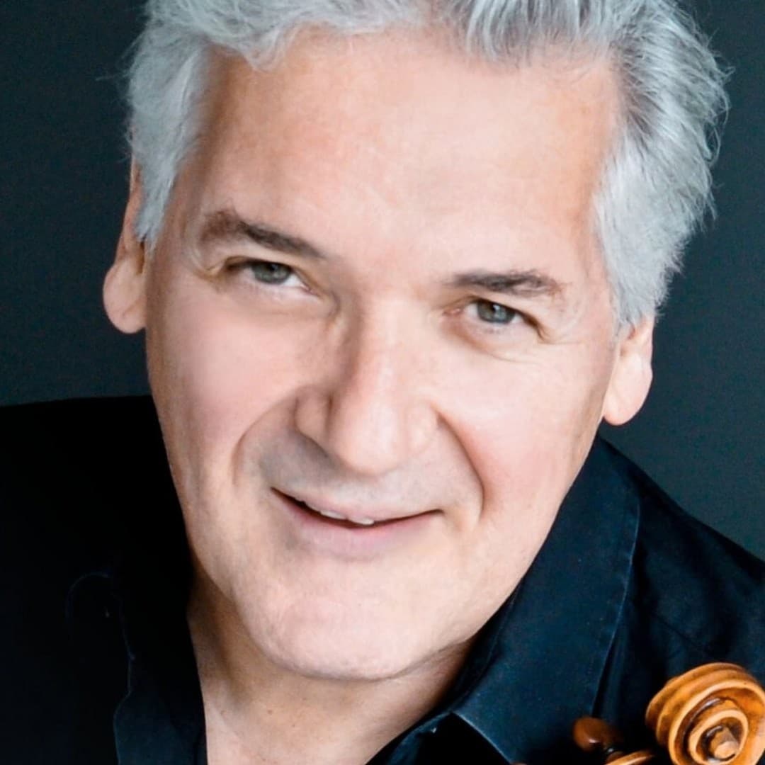 Person with gray hair smiles, wearing a black shirt. A violin's scroll is visible on the right shoulder. Dark background.