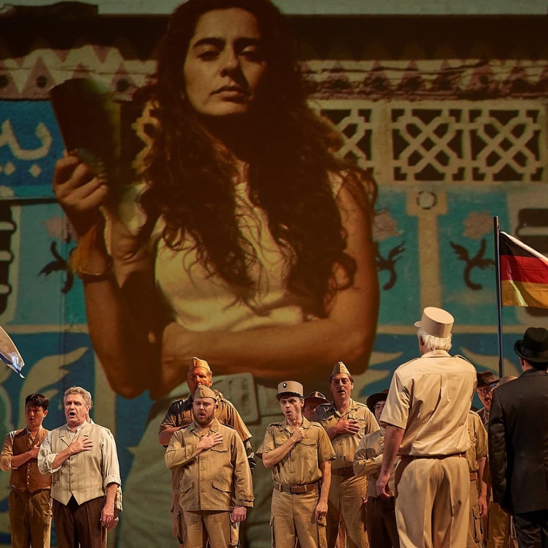 People in uniform and formal attire holding hands to chests, standing before projection of woman, two flags, blue and white, black, red, gold, text in