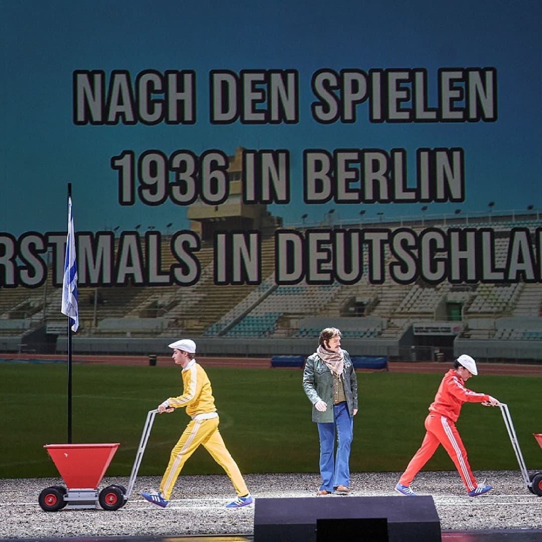 Two people with white hats push red carts on stage, one in yellow, one in red; person in casual clothes stands between them. Text reads: "NACH DEN SPIELEN 1936