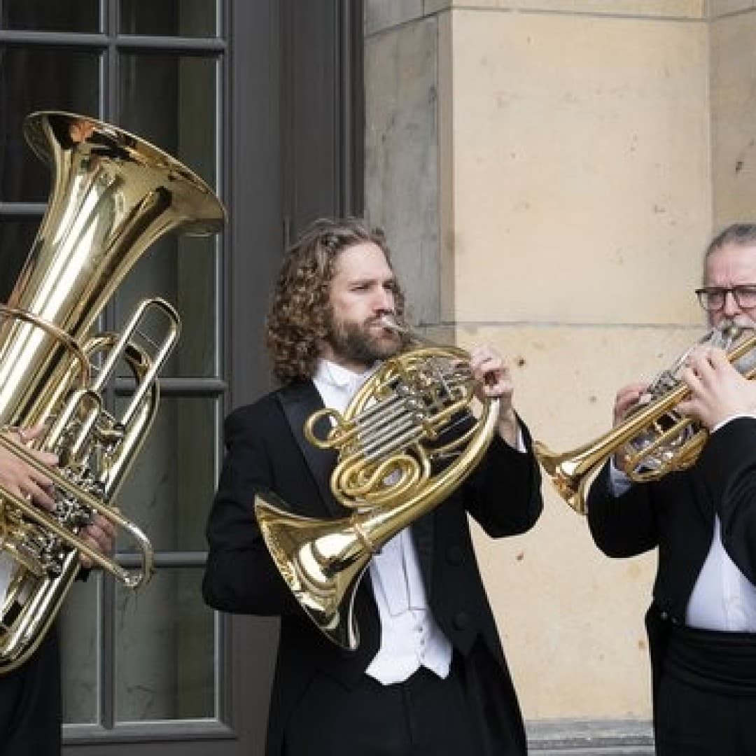 Five people in black suits play brass instruments outdoors: trombone, tuba, French horn, and two trumpets. They stand in front of large windows and a stone