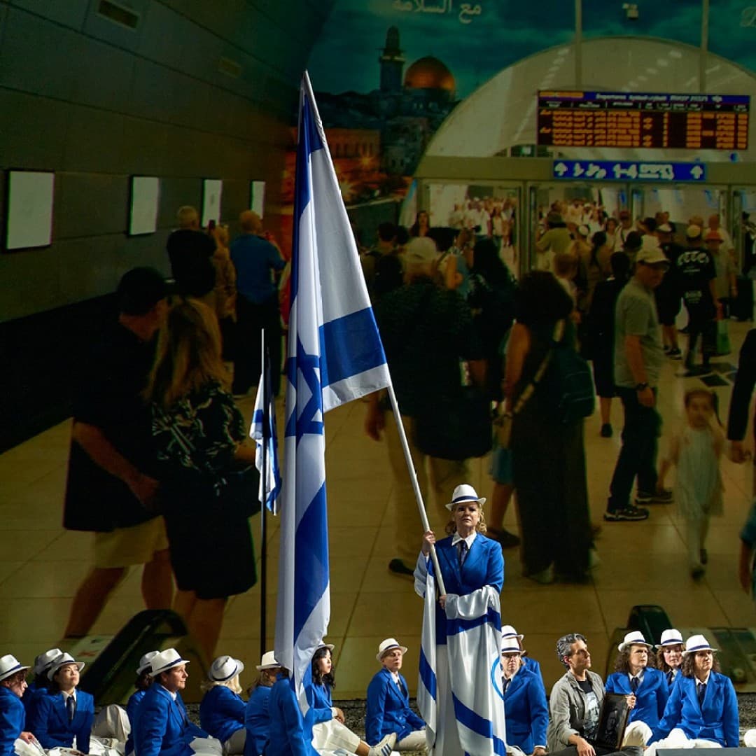 People in blue uniforms and hats sit and stand with an Israel flag in front of a wall-sized photo of a crowded corridor.